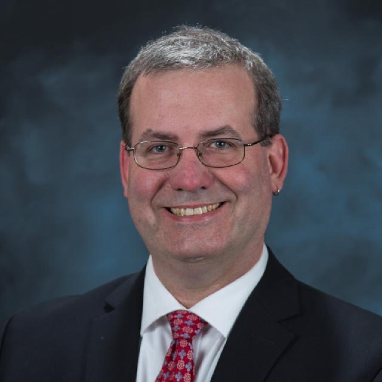 white male with glasses wearing dark jacket and red tie